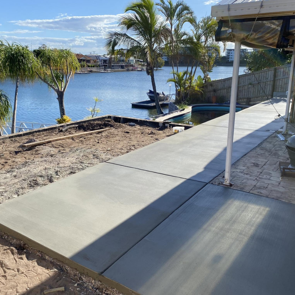 Wide concrete footpath poured in a waterfront backyard overlooking a canal with palm trees and a moored boat